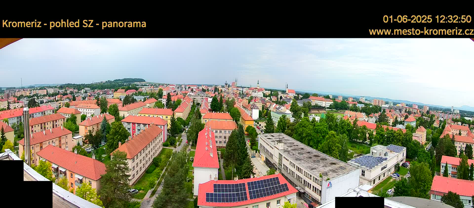 A panoramic view of a town on an overcast day, showing numerous buildings with red tile roofs, interspersed with green trees and patches of grass, stretching towards a distant horizon.