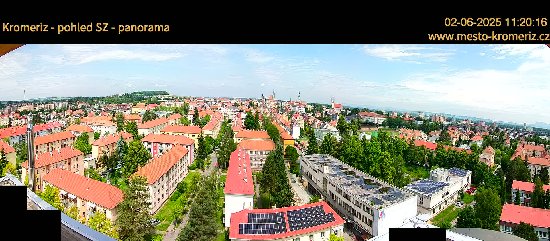 A panoramic view of a town on a partly cloudy day, showing many buildings with red roofs, interspersed with green trees, and a distant, rolling landscape.