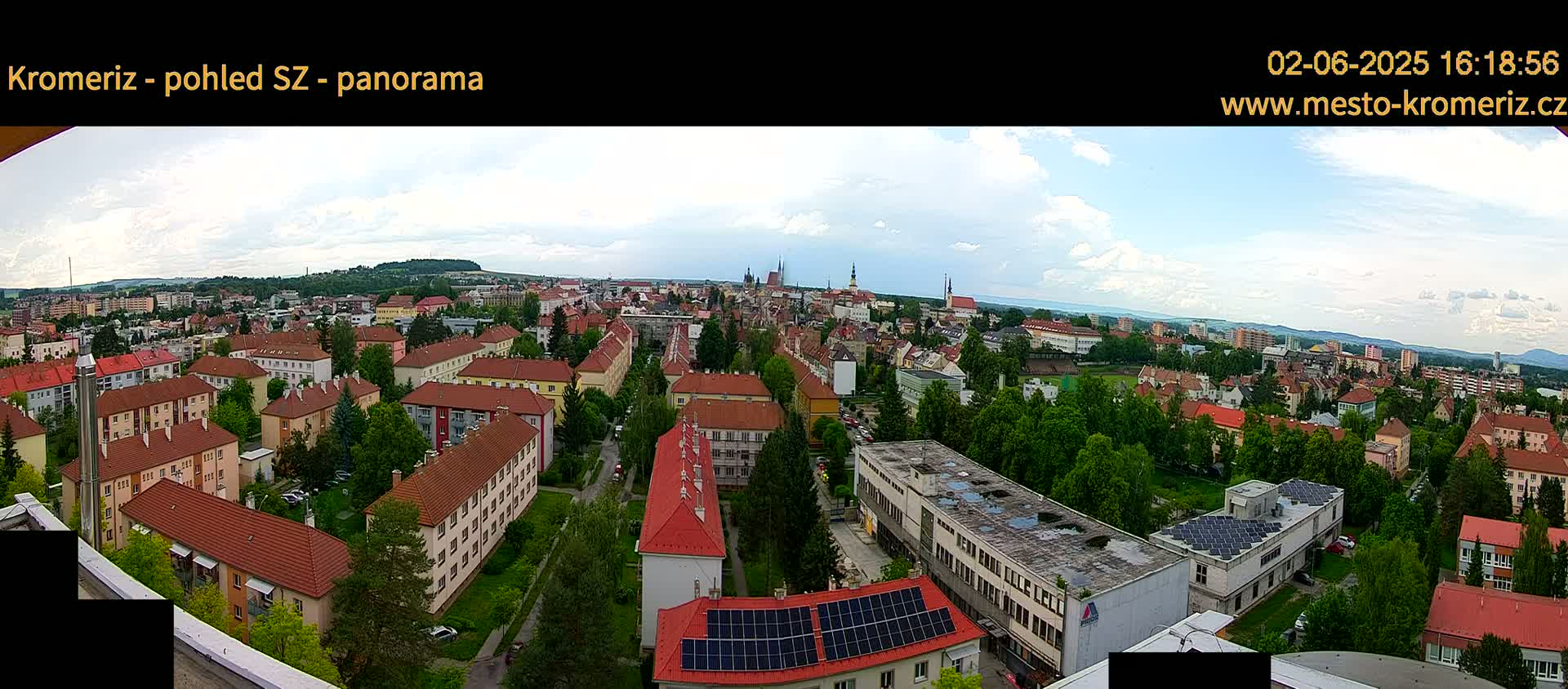 A panoramic view of a town under a partly cloudy sky shows numerous buildings with red tile roofs, interspersed with green trees, and a distant horizon of hills.