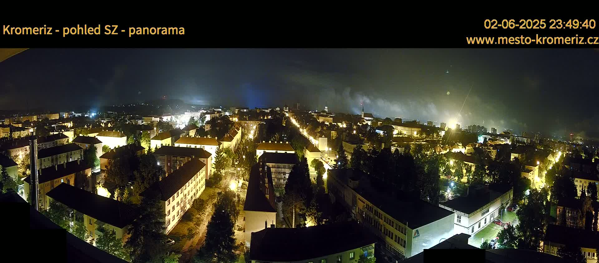 A nighttime panoramic view of a town, illuminated by streetlights, with a hazy sky and distant lights possibly from fireworks.