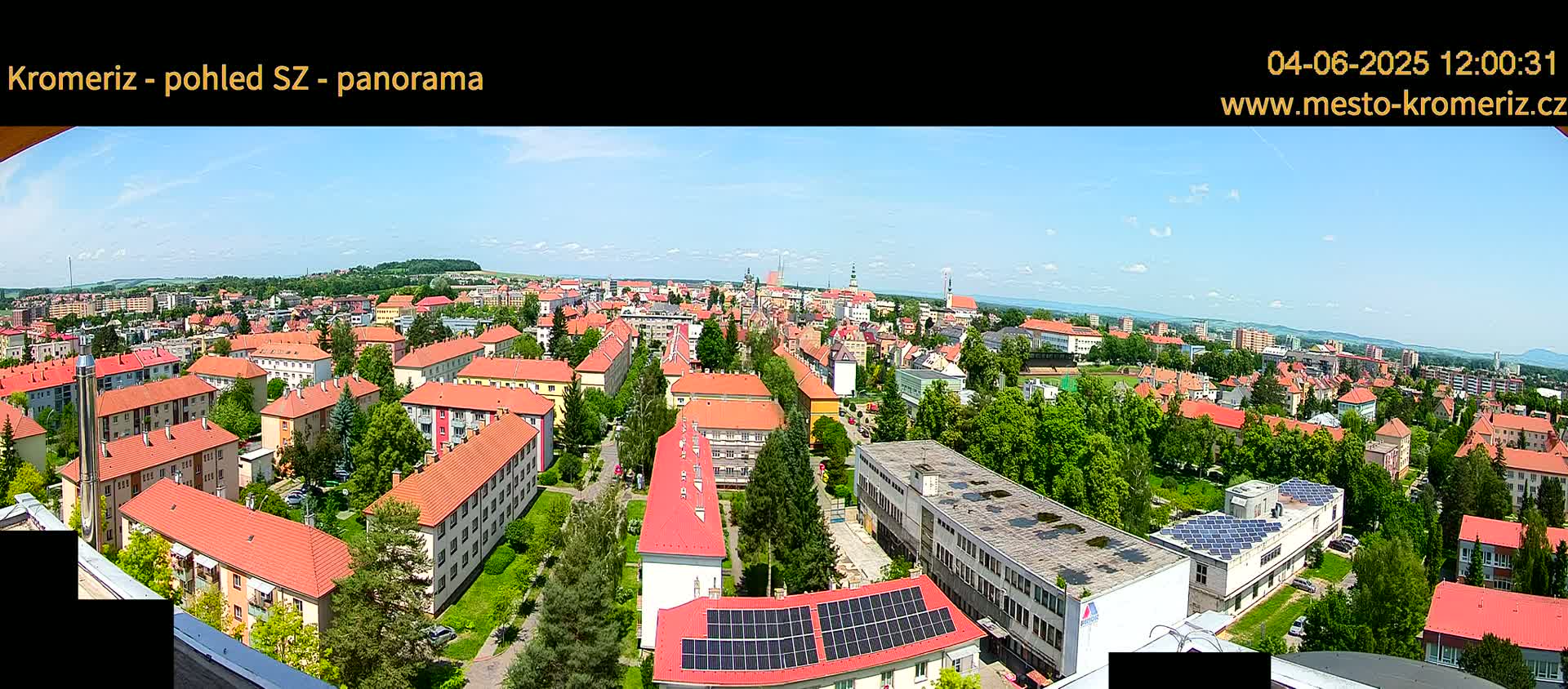 A panoramic view of a town on a sunny day shows many buildings with red tile roofs, surrounded by green trees and landscaping.