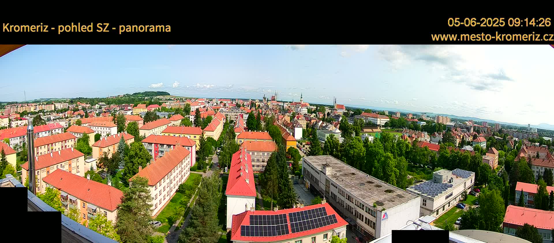 A panoramic view of a town under a mostly clear sky shows many buildings with red-tiled roofs, surrounded by green trees.