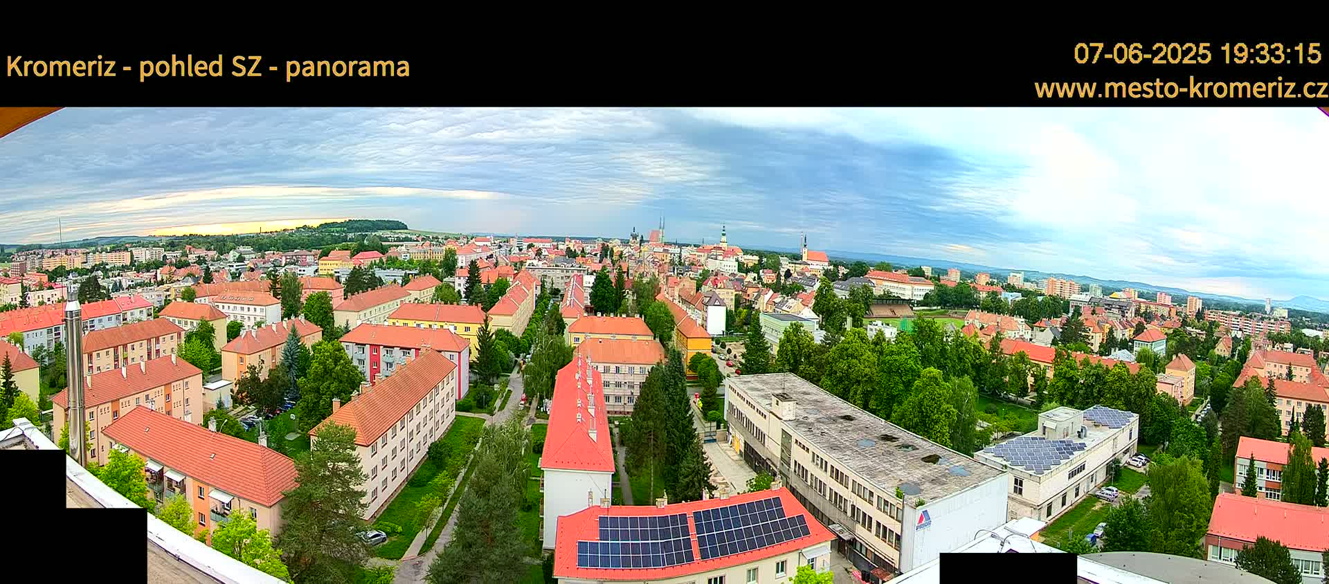 A panoramic view of a town under a partly cloudy sky shows many low-rise buildings with red tile roofs, interspersed with green trees and some larger buildings.