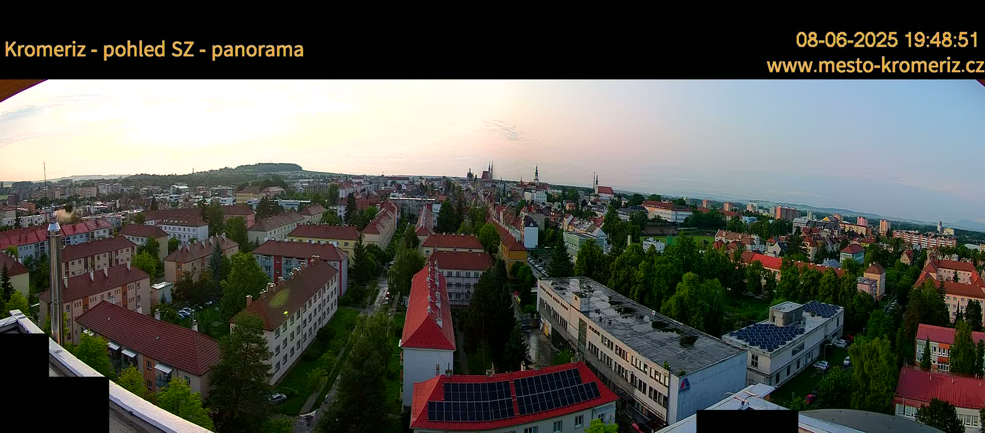 An expansive view of a city at sunset, showing numerous buildings with red roofs and lush green trees interspersed.