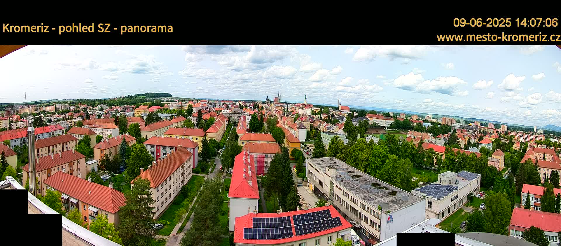 A panoramic view of a town under a partly cloudy sky shows many buildings with red-tiled roofs, green trees, and a large building with solar panels on its roof.