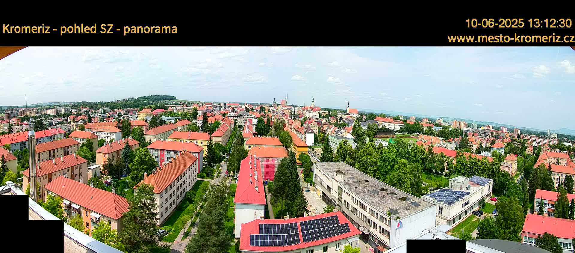 A panoramic view of a town with predominantly red-roofed buildings under a partly cloudy sky shows a mix of residential and commercial structures, including buildings with solar panels, surrounded by green trees.