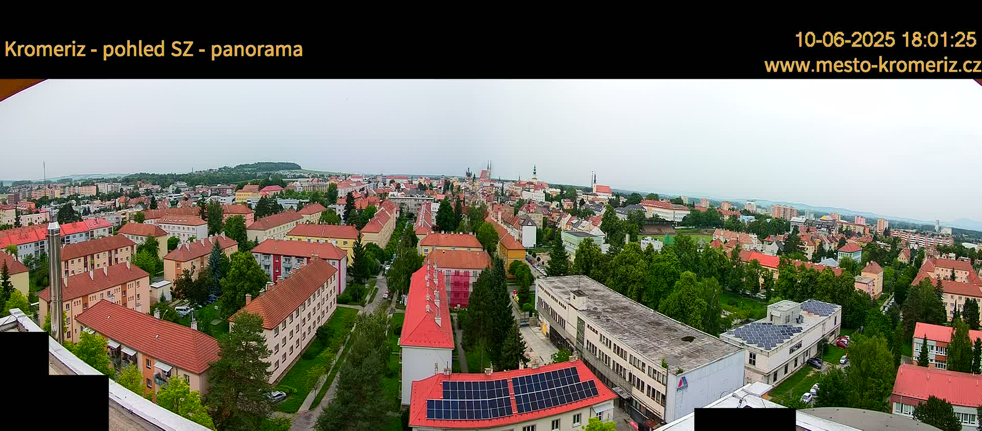 A panoramic view of a town shows many red-roofed buildings and green trees under an overcast sky.