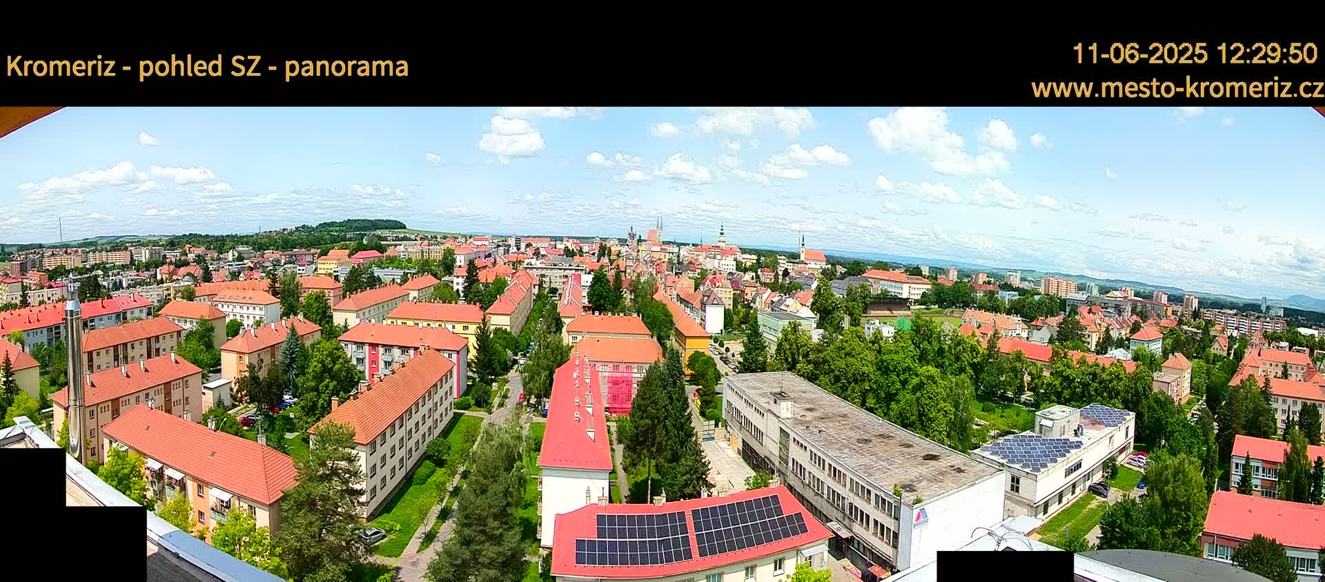 A panoramic view of a town on a partly sunny day shows many buildings with red roofs and green trees, extending to a distant horizon.