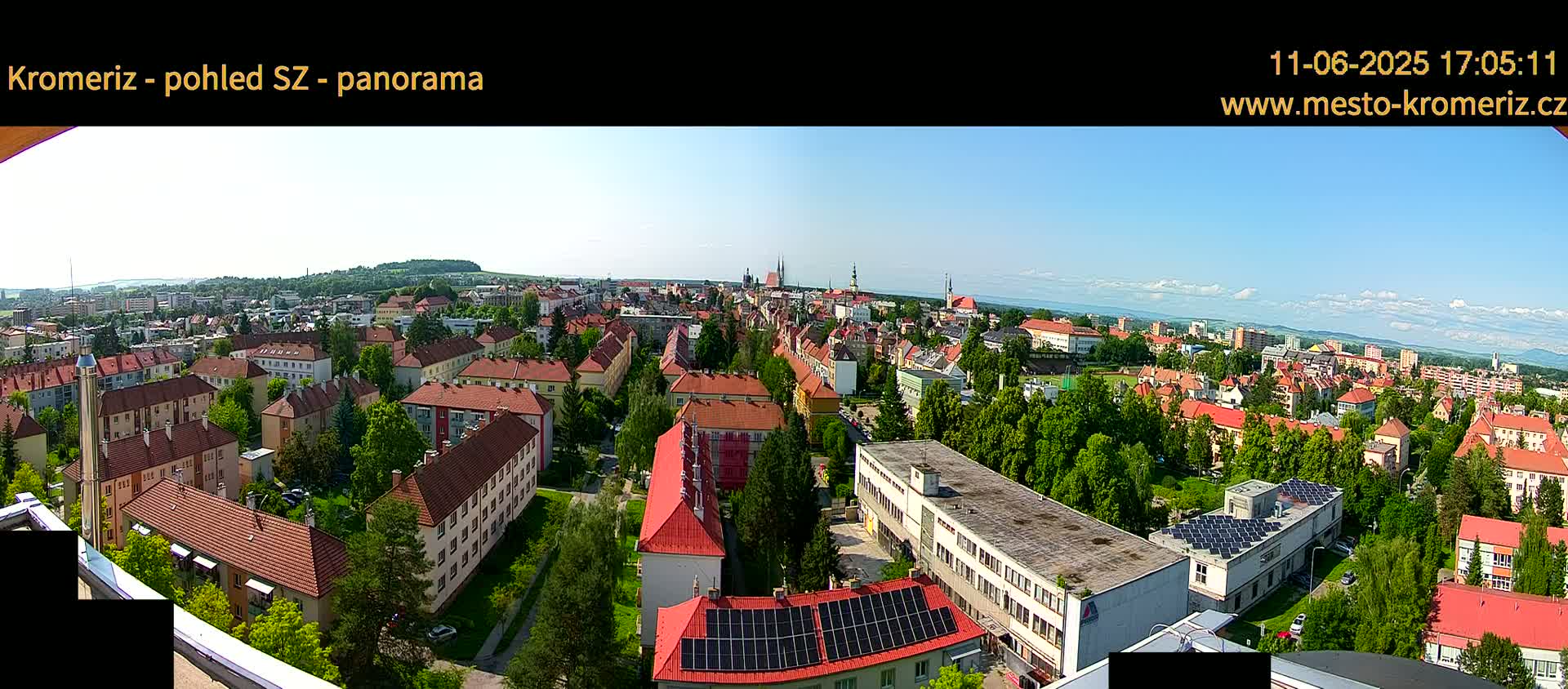 A panoramic view of a town on a sunny day shows many buildings with red-tiled roofs, surrounded by green trees and a clear blue sky with a few clouds.