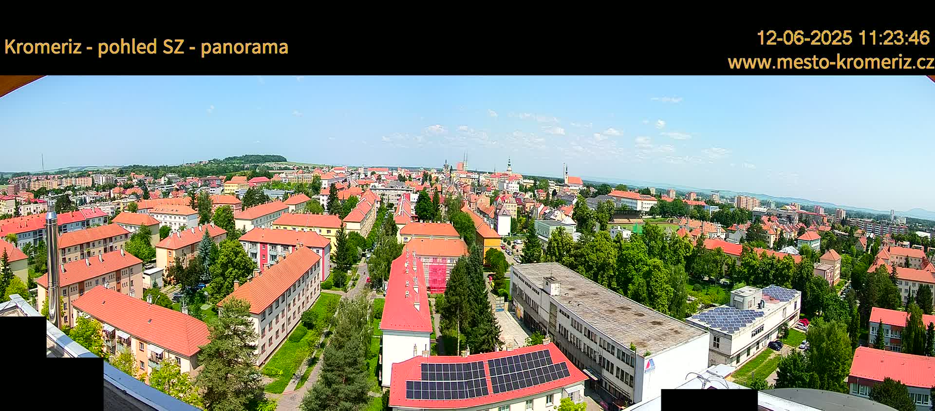 A panoramic view of a town on a sunny day shows many buildings with red-tiled roofs, interspersed with green trees and patches of grass.