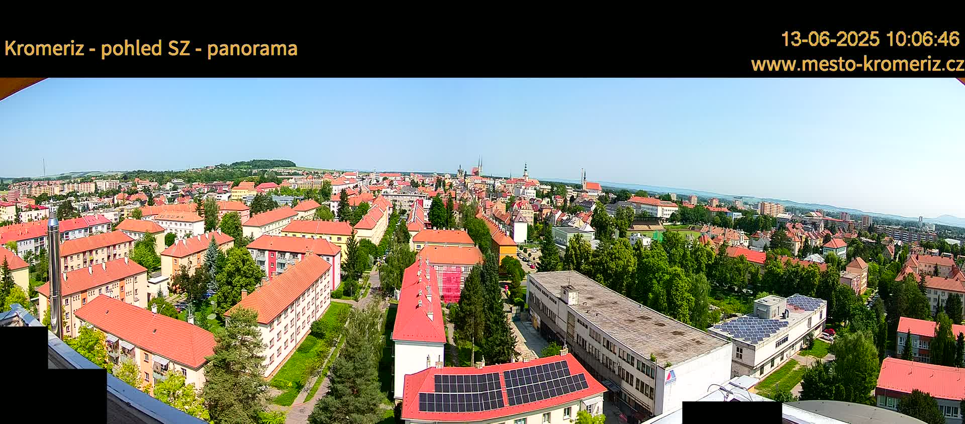 A panoramic view of a town on a sunny day shows many red-roofed buildings surrounded by lush green trees.