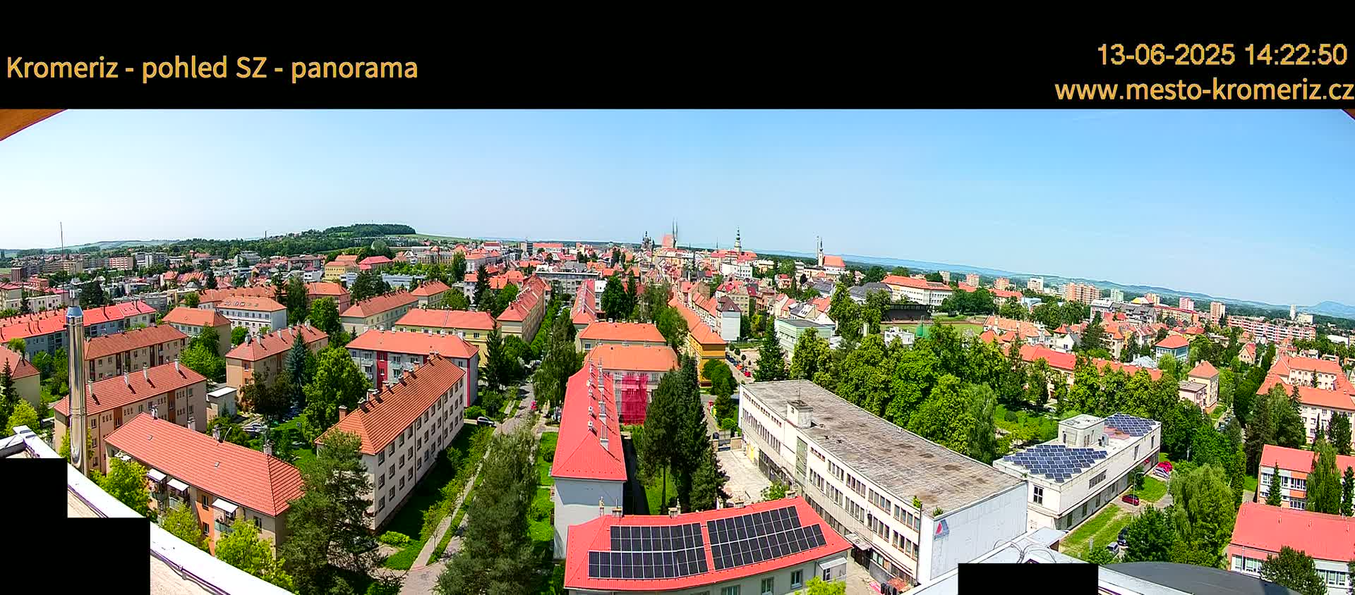 A panoramic view of a town on a sunny day shows many red-roofed buildings, interspersed with green trees, extending to a distant horizon.