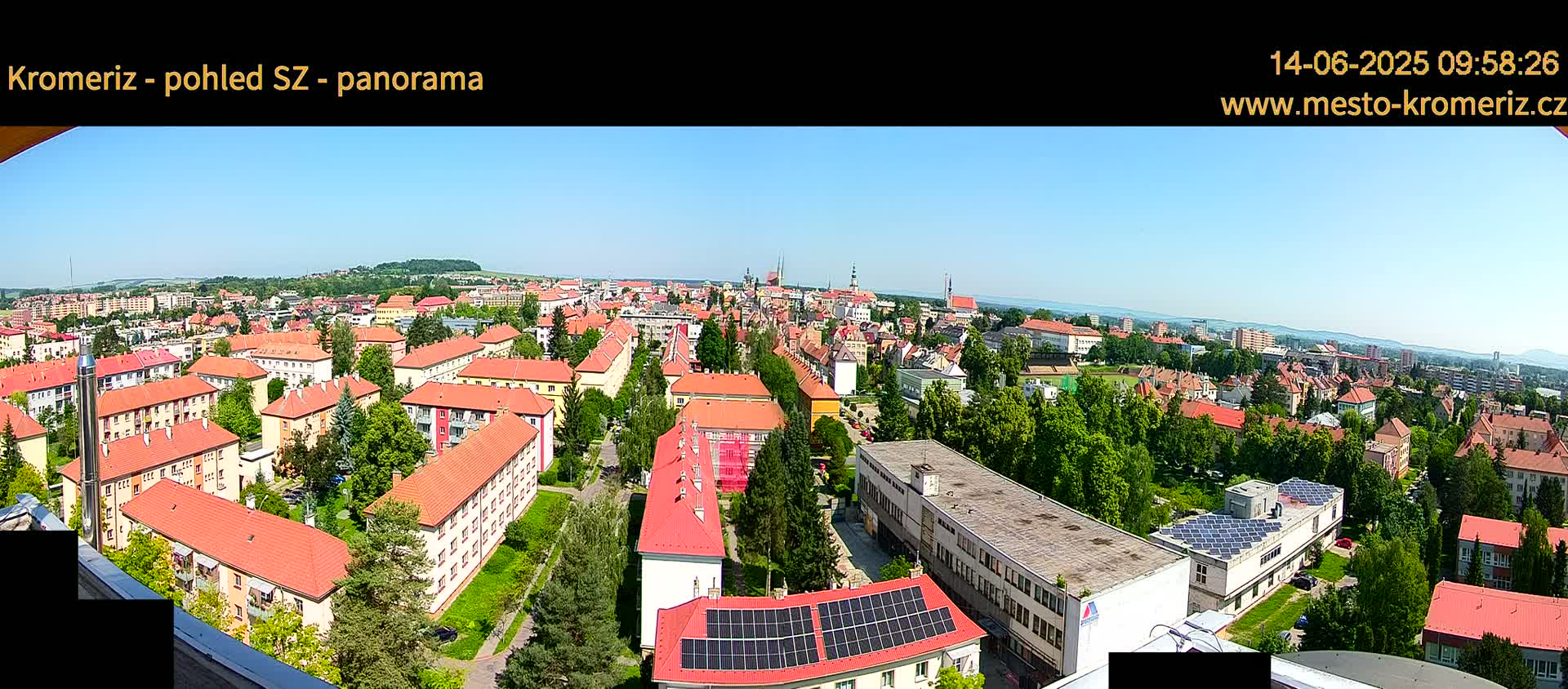 A panoramic view of a town on a sunny day shows numerous buildings with red-tiled roofs, interspersed with green trees and vegetation.