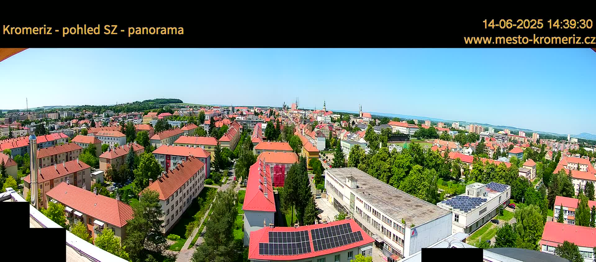 A panoramic view on a sunny day shows a town with many low-rise buildings, mostly with red-tiled roofs, surrounded by green trees and vegetation.