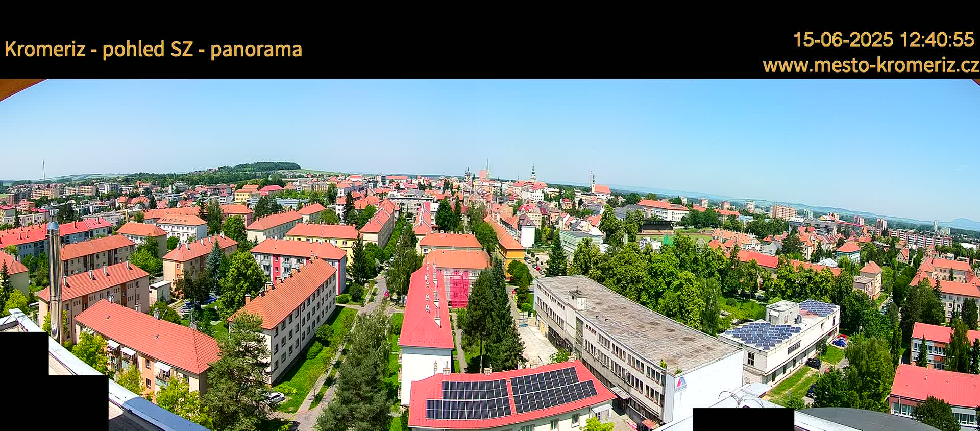 A panoramic view of a town on a clear, sunny day shows a dense collection of buildings with red-tiled roofs, interspersed with green spaces and trees.