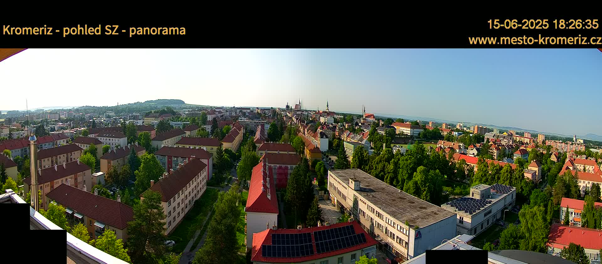 A panoramic view of a town on a clear, sunny day shows many low-rise buildings surrounded by lush green trees.