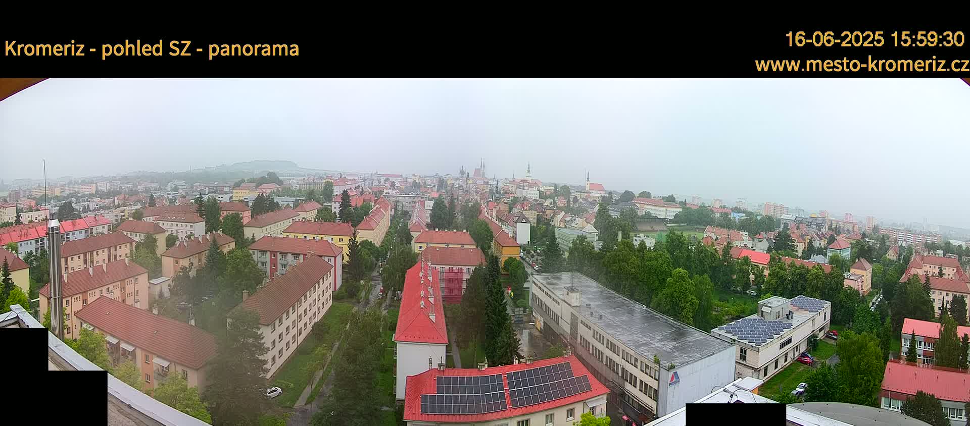 A panoramic view of a town on an overcast day, showing numerous buildings, trees, and a large expanse of greenery.