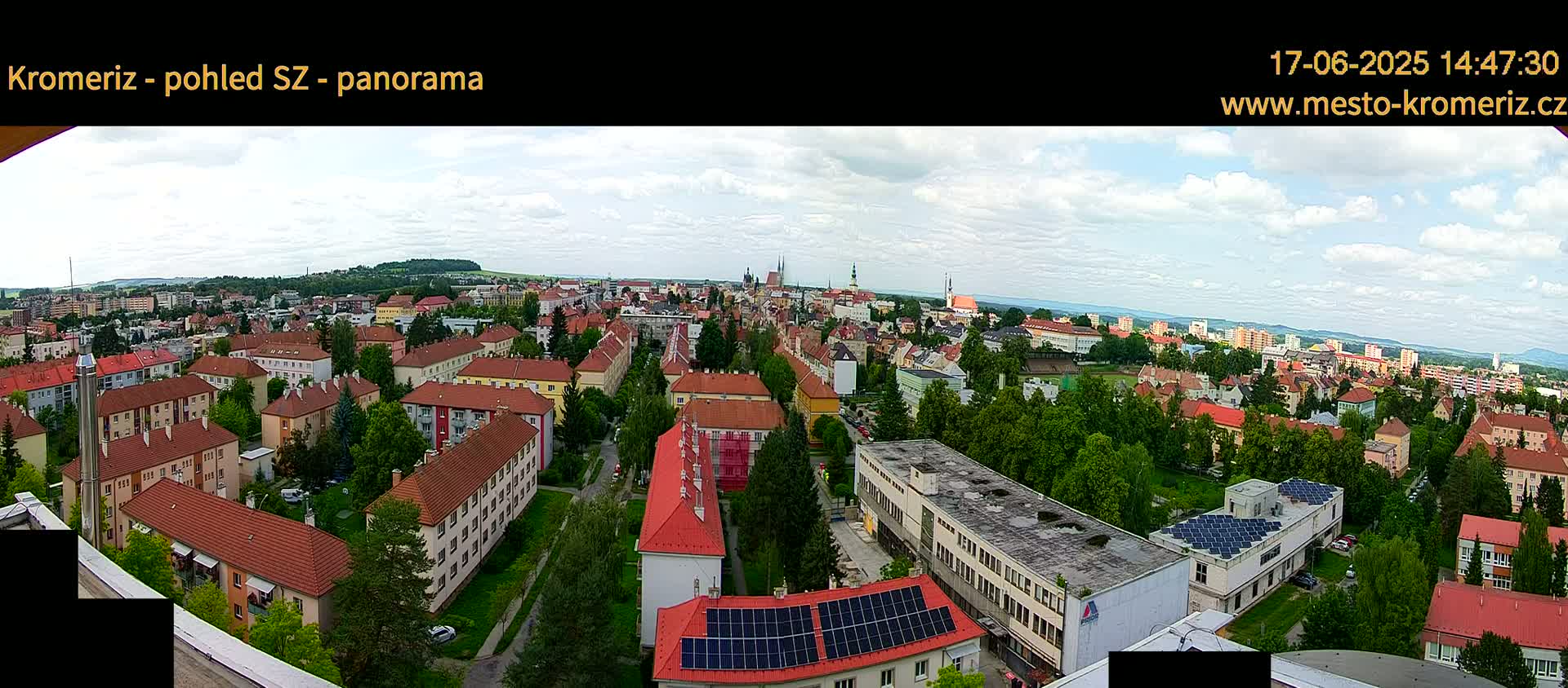 A panoramic view of a town on a partly cloudy day, showing many red-roofed buildings, green trees, and a large building with solar panels.
