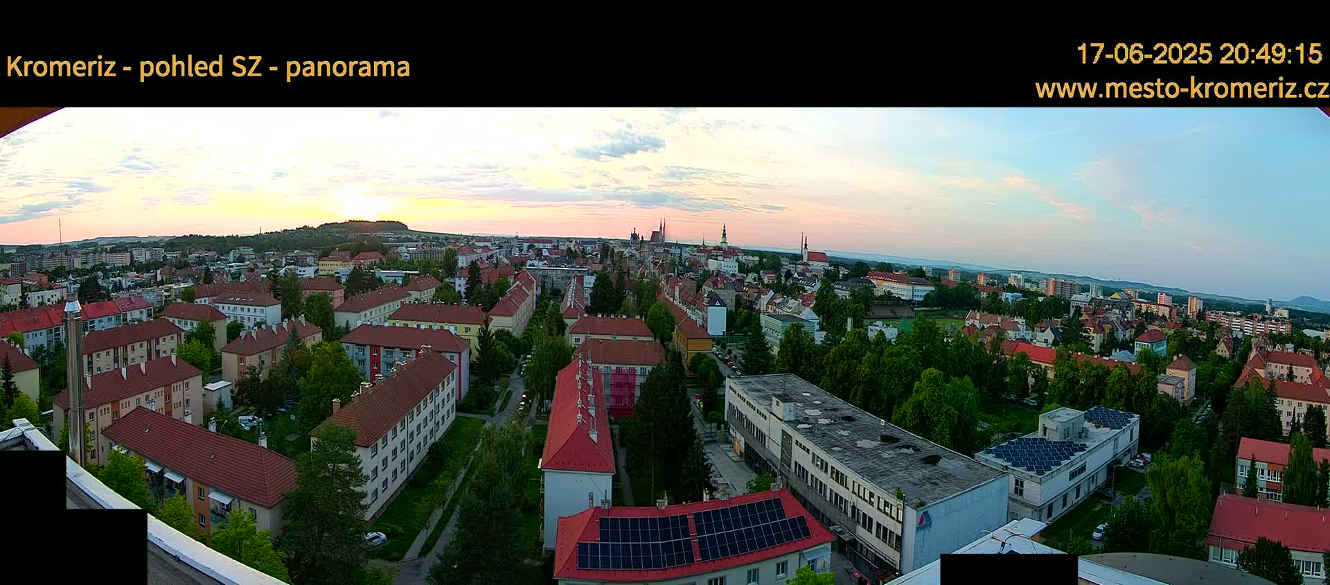 An aerial view of a European town at sunset, showing a mix of residential buildings and greenery under a mostly clear sky.