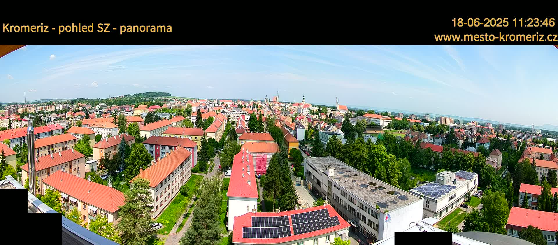 A panoramic view of a town with many red-roofed buildings under a mostly sunny sky shows lush green trees and distant hills.