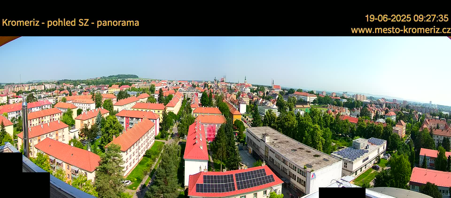 A panoramic view of a town on a sunny day, showing numerous red-roofed buildings interspersed with green trees and several buildings with solar panels on their roofs.