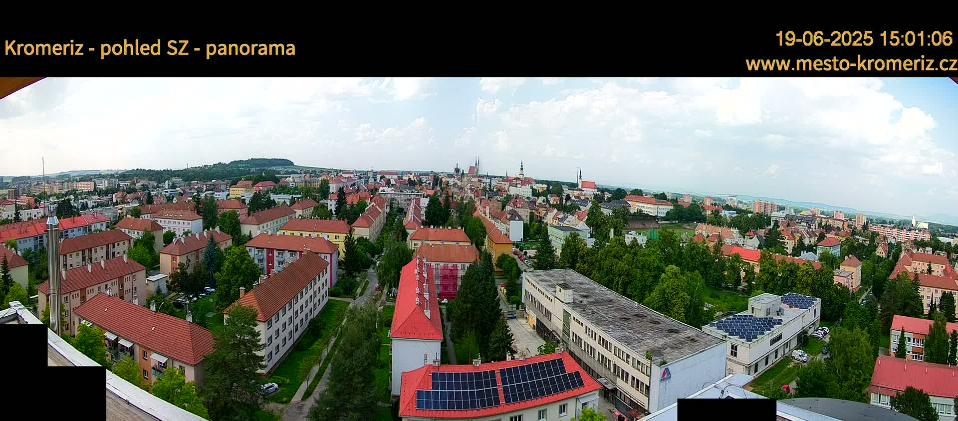 A panoramic view of a town on a partly cloudy day shows many buildings with red roofs, interspersed with green trees, and a large building with solar panels on the roof in the foreground.