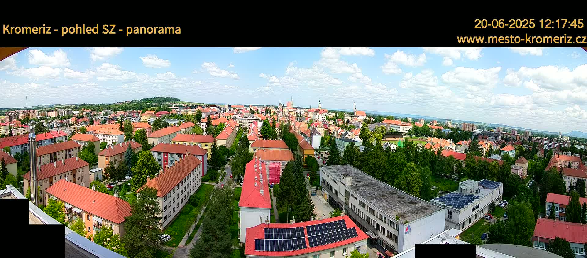 A panoramic view of a town on a partly cloudy day shows many red-roofed buildings surrounded by green trees.