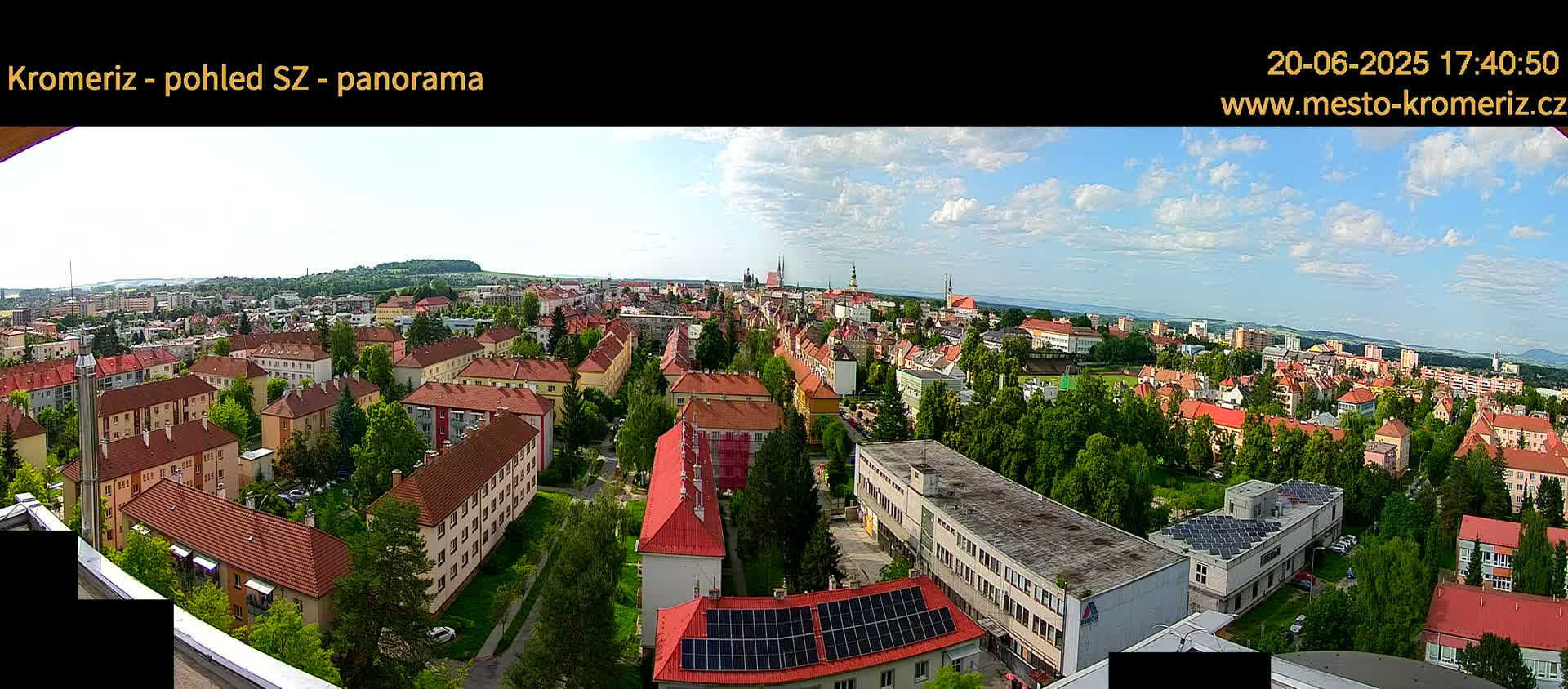 A panoramic view of a town on a partly cloudy day shows many red-roofed buildings interspersed with green trees, with a larger, flatter building with solar panels in the foreground.