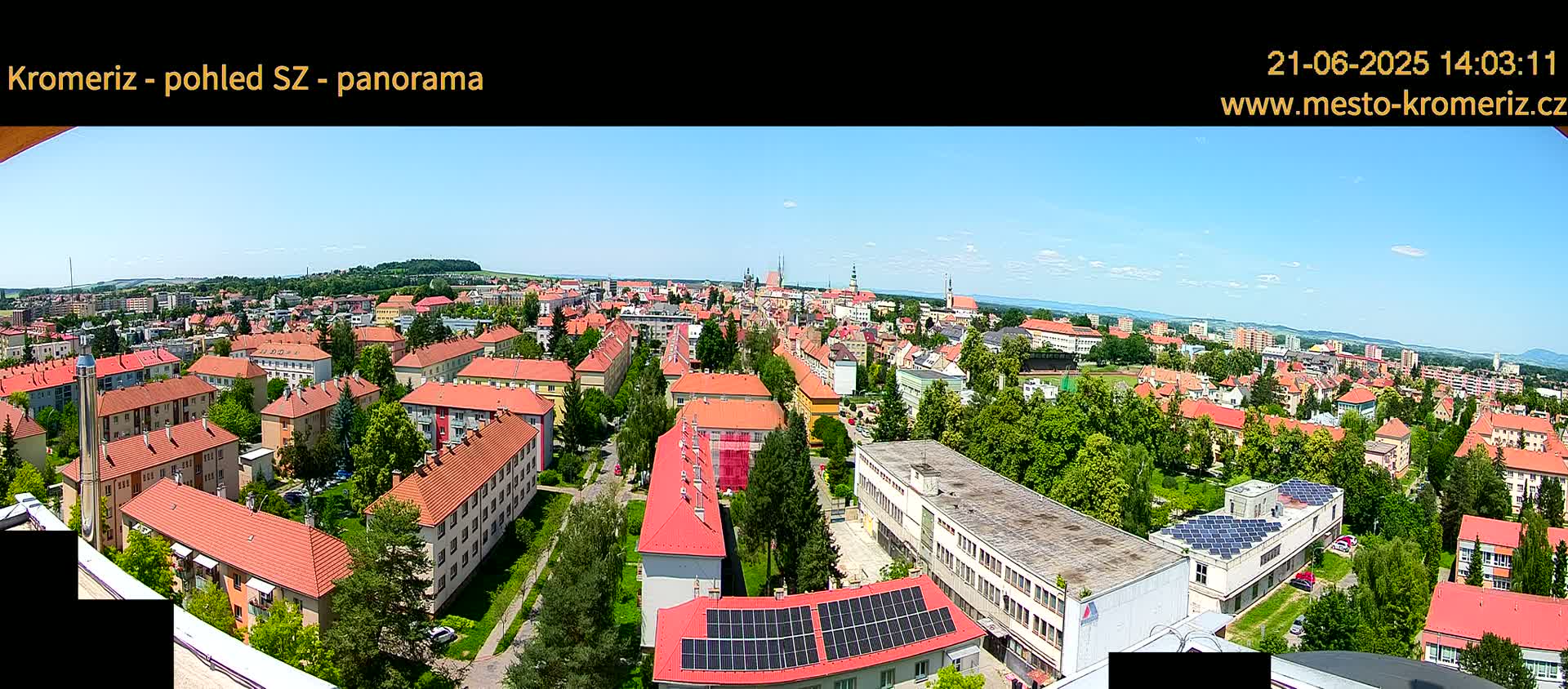 A panoramic view of a town on a sunny day, showing many low-rise buildings with red tile roofs, surrounded by green trees and landscaping.
