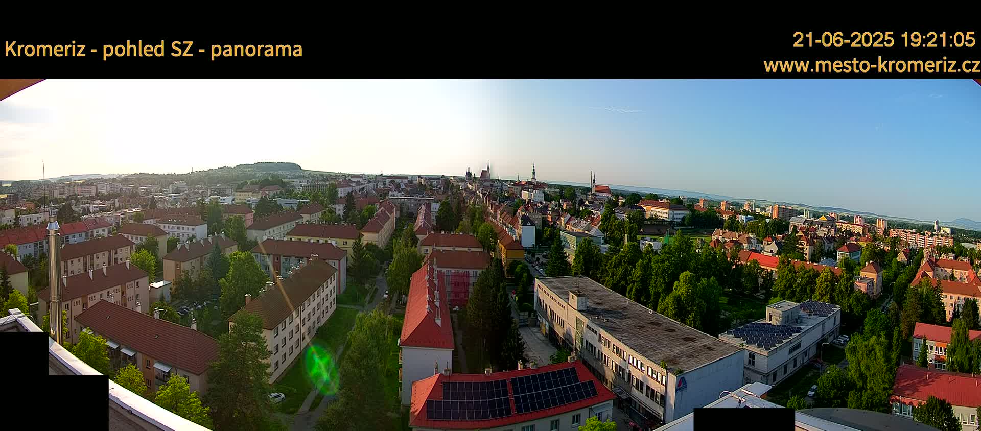 A panoramic view of a town under a clear, bright sky shows many buildings with red-tiled roofs, surrounded by lush green trees.