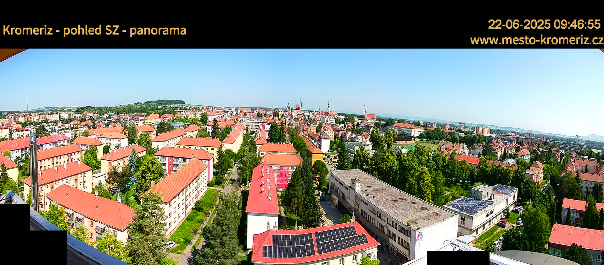 A panoramic view of a town on a sunny day shows many buildings with red roofs, interspersed with green trees and vegetation.