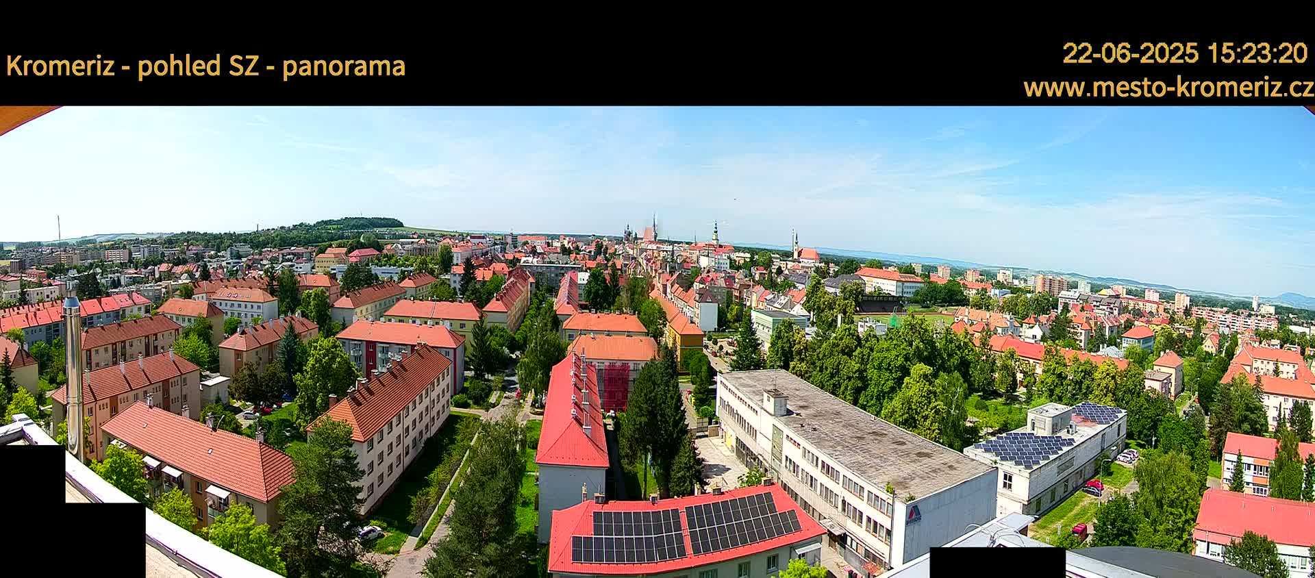 An aerial view shows a town of predominantly red-roofed buildings under a clear, sunny sky.
