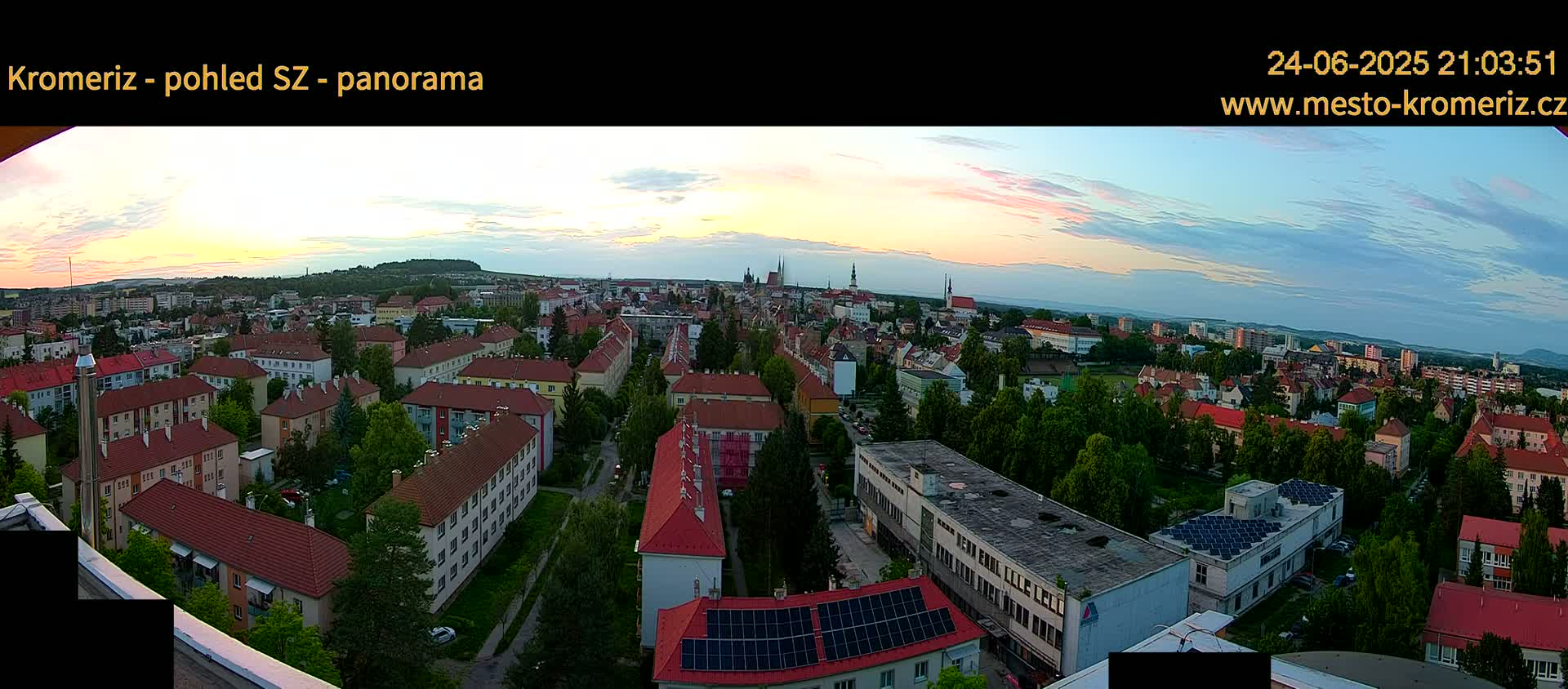 A panoramic view of a town at sunset, showing many red-roofed buildings and lush green trees under a partly cloudy sky.