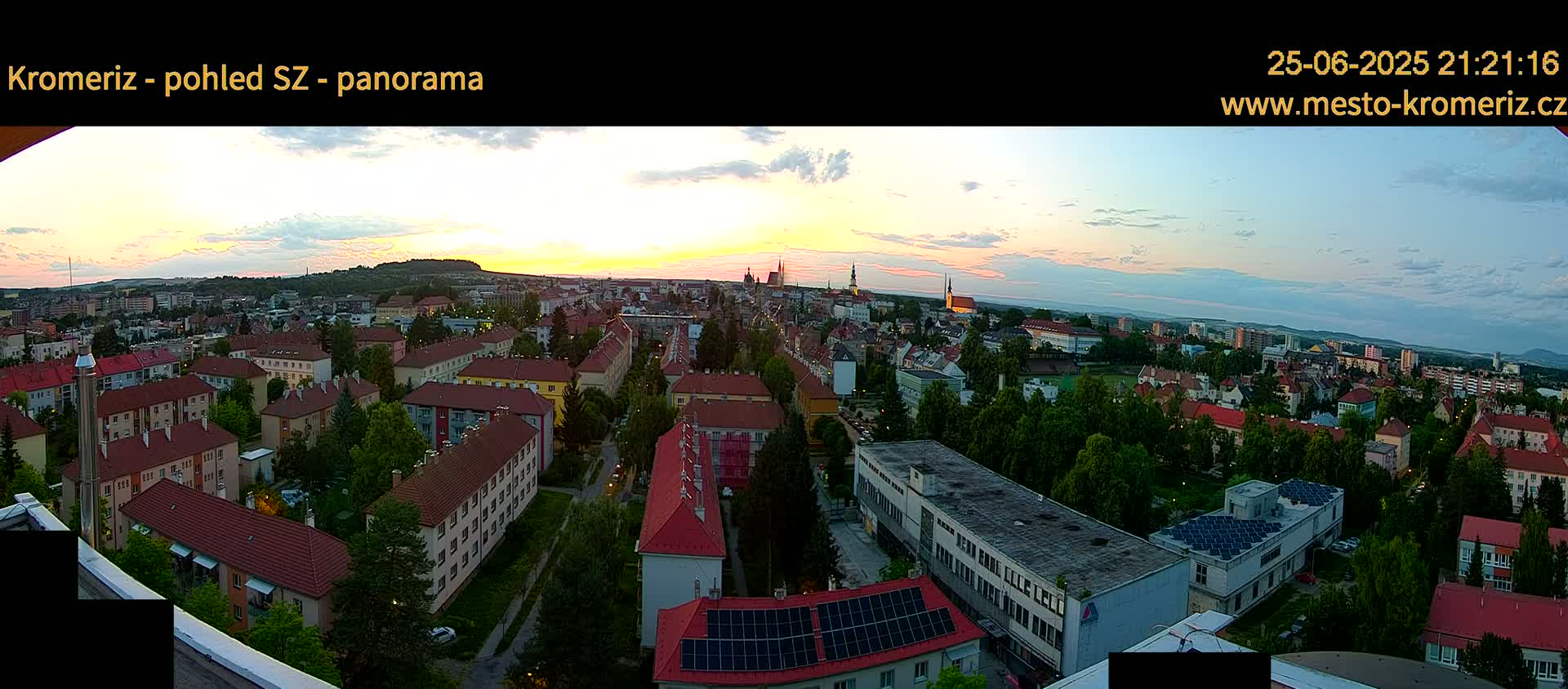 A panoramic view of a town at sunset, showing many residential buildings and trees under a partly cloudy sky.