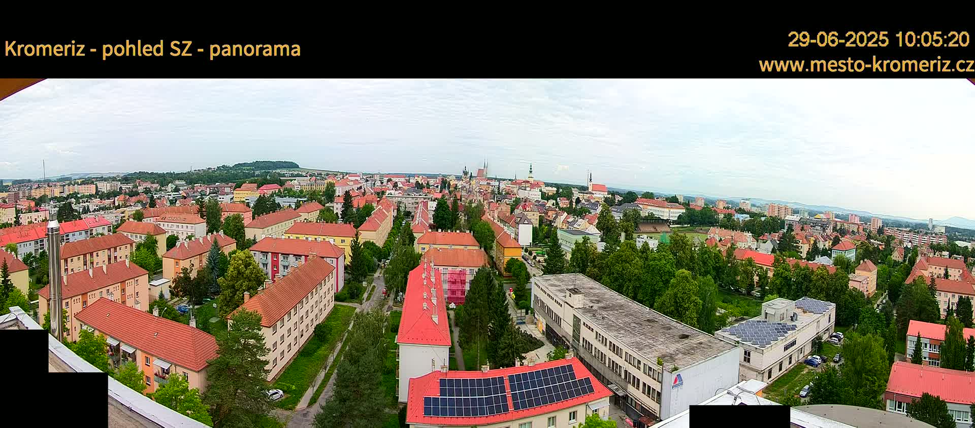 A panoramic view of a town on an overcast day, showing many low-rise buildings with red-tiled roofs, interspersed with green trees and surrounded by hills in the distance.