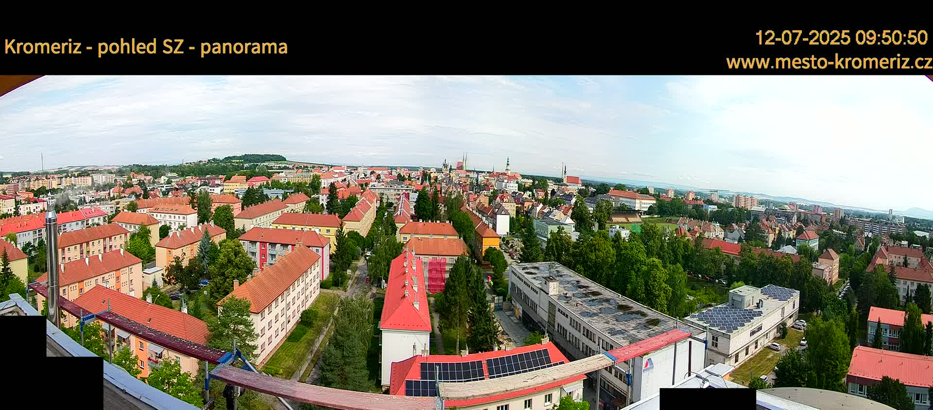 A panoramic view of a town under partly cloudy skies shows numerous red-roofed buildings, interspersed with green trees, stretching to the horizon.