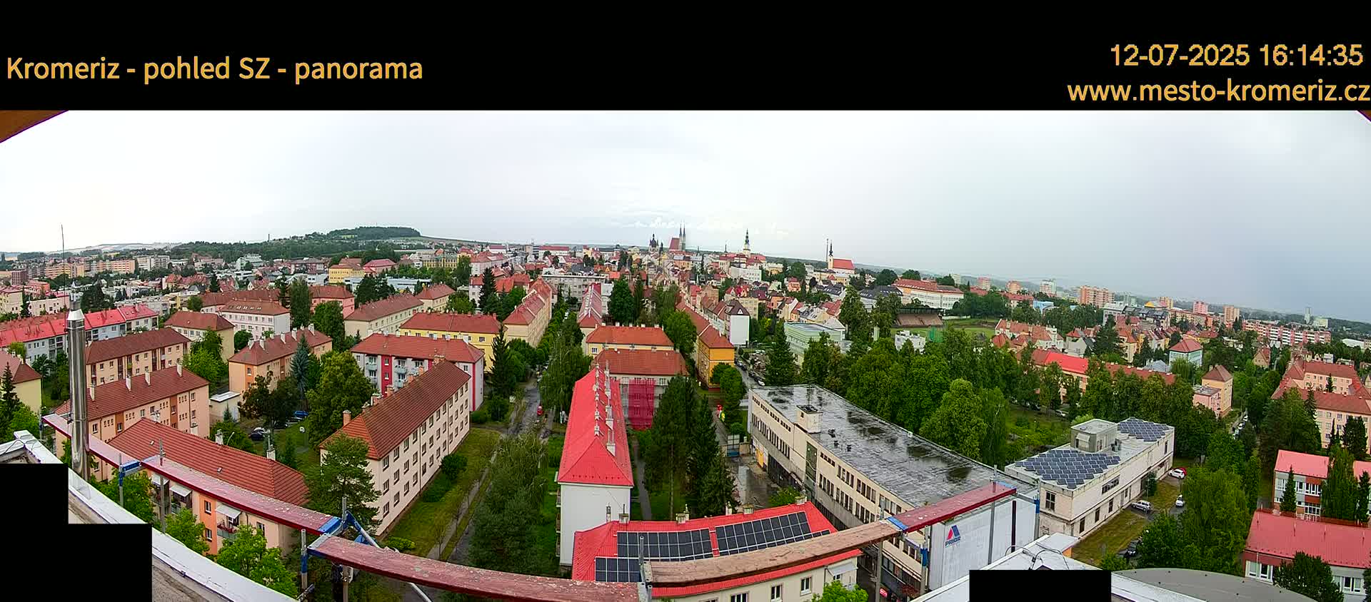 A panoramic view of a town under partly cloudy skies shows numerous red-roofed buildings, interspersed with green trees, stretching to the horizon.