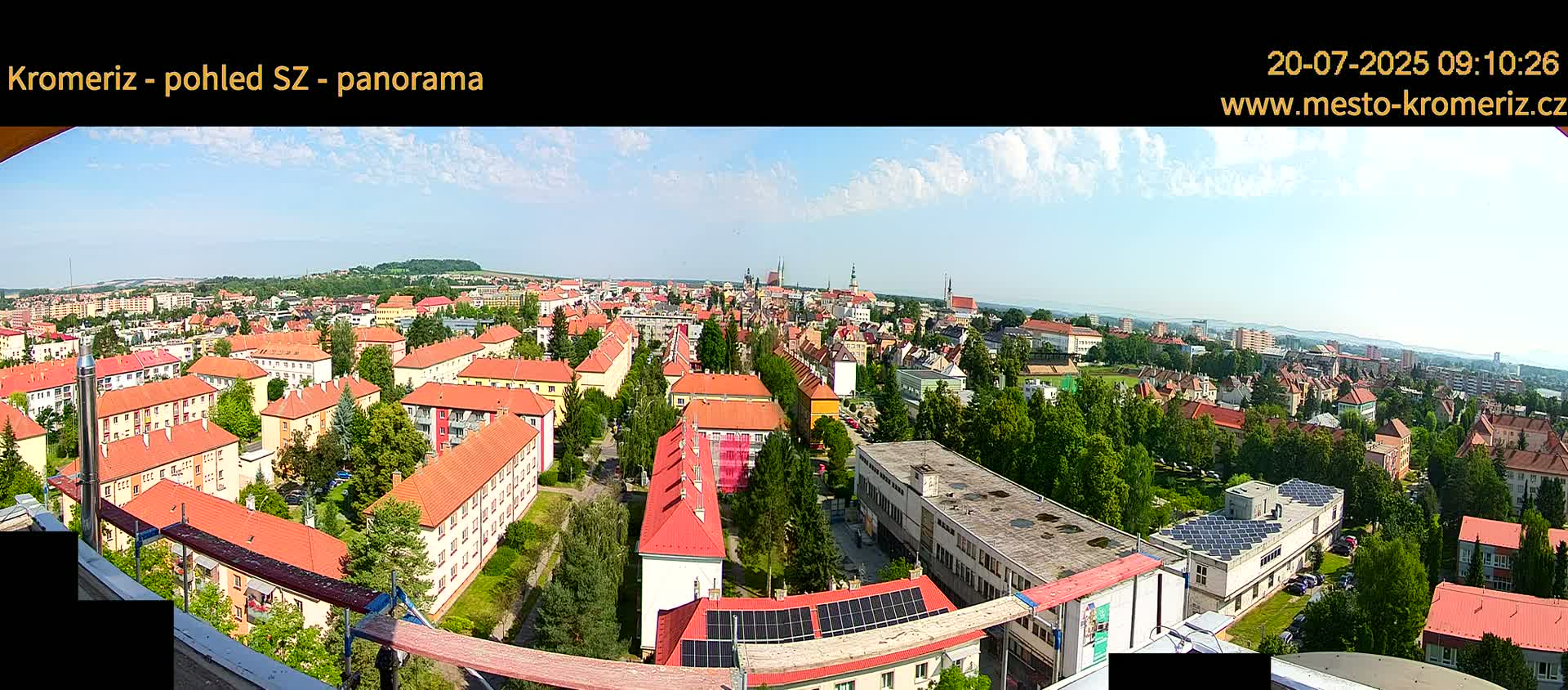 A panoramic view on a sunny day shows a sprawling town with predominantly red-roofed buildings, interspersed with green trees, and a distant horizon of hills.