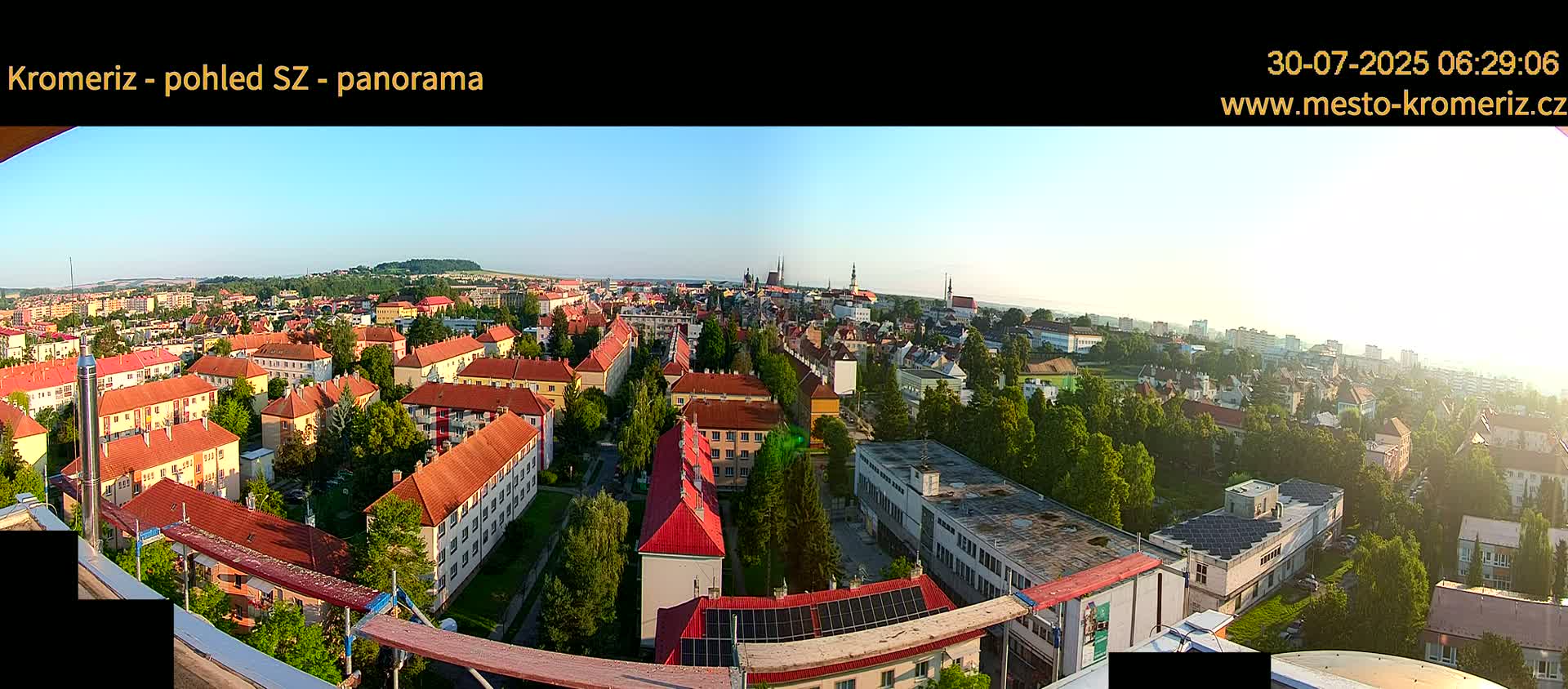 A panoramic view on a sunny day shows a sprawling town with predominantly red-roofed buildings, interspersed with green trees, and a distant horizon of hills.