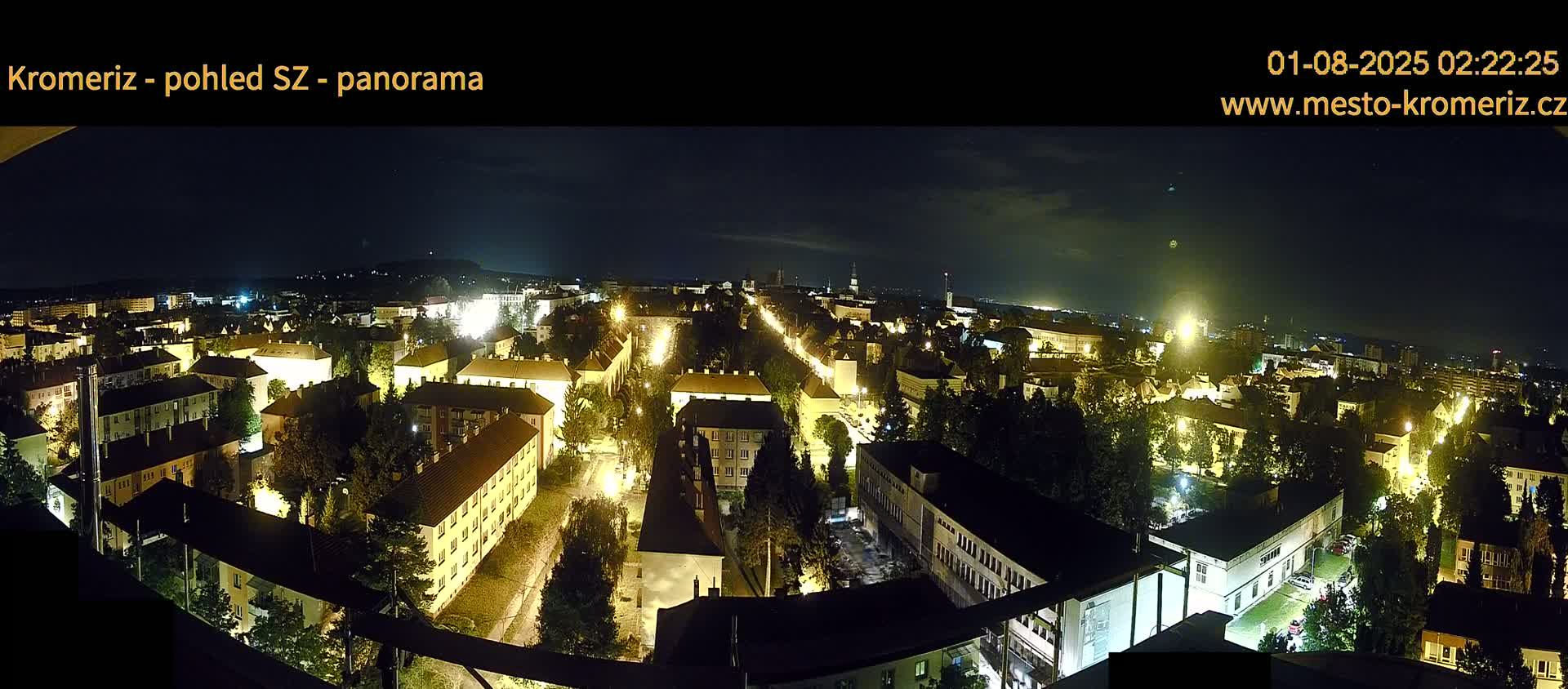 A nighttime panoramic view of a city with streetlights illuminating buildings and trees under a partly cloudy sky.