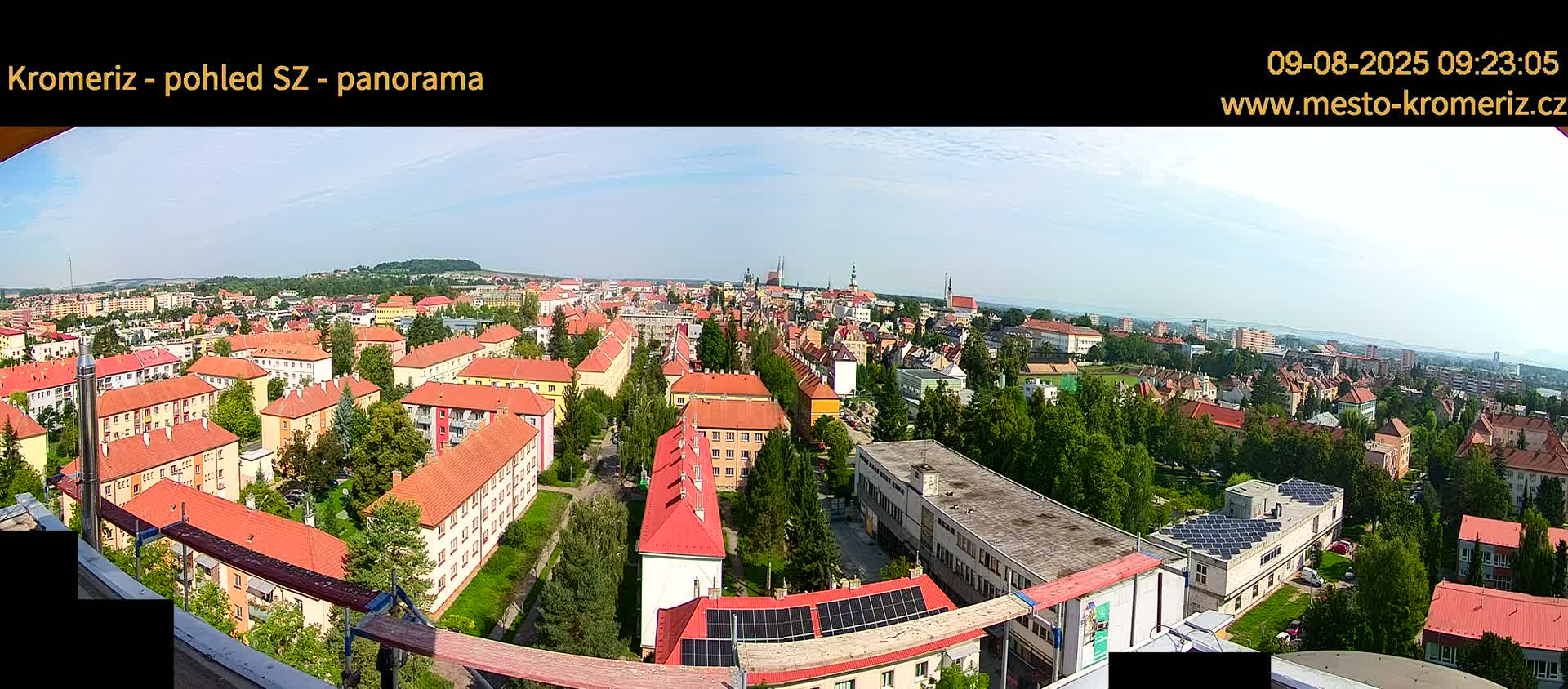 A panoramic view on a sunny day shows a town with many red-roofed buildings, green trees, and a distant hilly landscape.