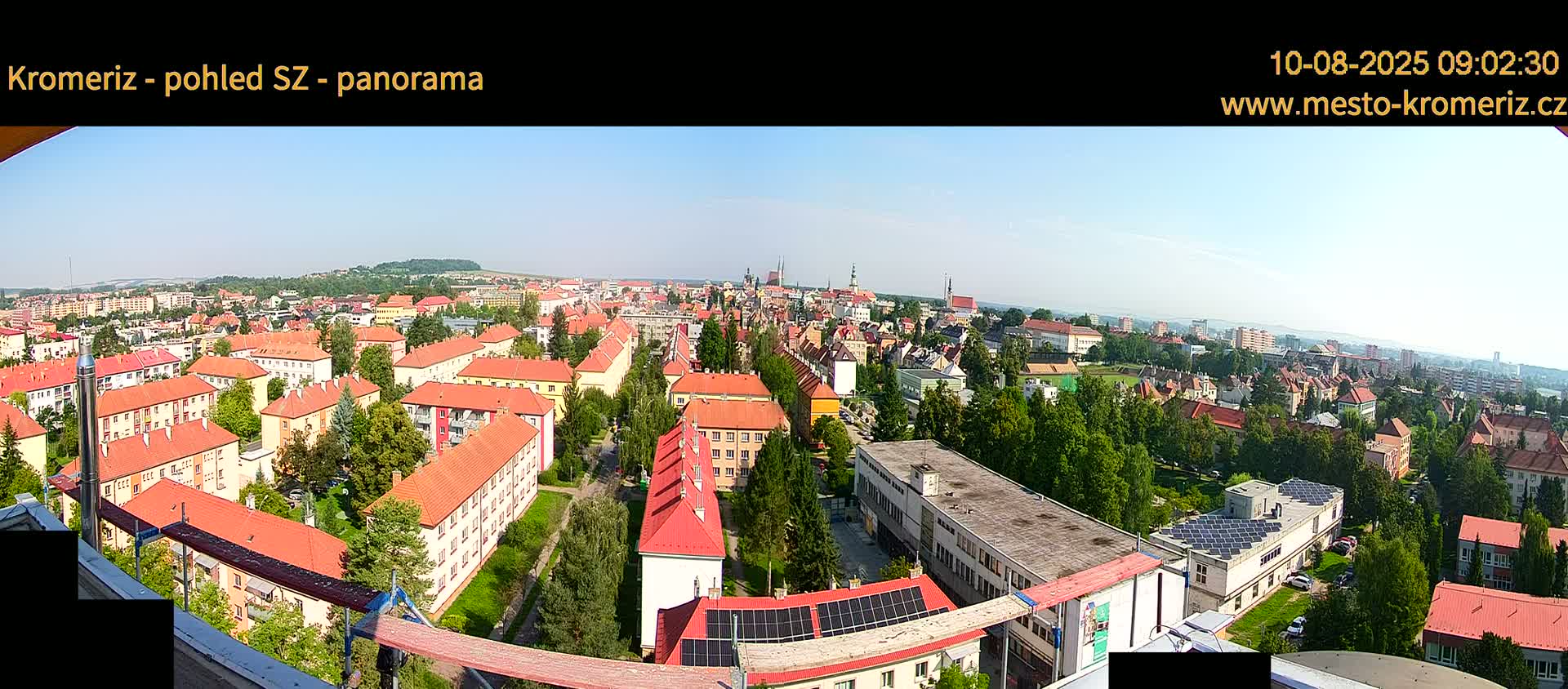 A panoramic view on a clear, sunny day shows a large town with many low buildings with red roofs, interspersed with green trees, extending to a horizon of more distant buildings and hills.