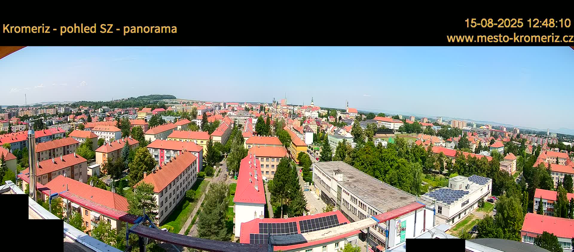 A panoramic view on a clear, sunny day shows a large town with many low buildings with red roofs, interspersed with green trees, extending to a horizon of more distant buildings and hills.