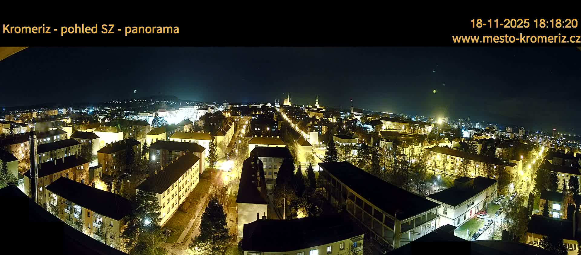 A panoramic view on a clear, sunny day shows a large town with many low buildings with red roofs, interspersed with green trees, extending to a horizon of more distant buildings and hills.
