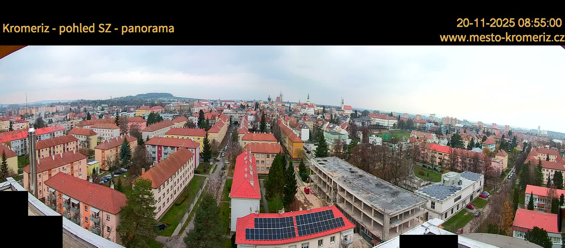 A sprawling panoramic view of a European town features dense residential buildings with predominantly red-tiled roofs, interspersed with bare trees and streets, a building with solar panels, and a large concrete structure in the foreground, all under a uniformly grey, overcast sky.