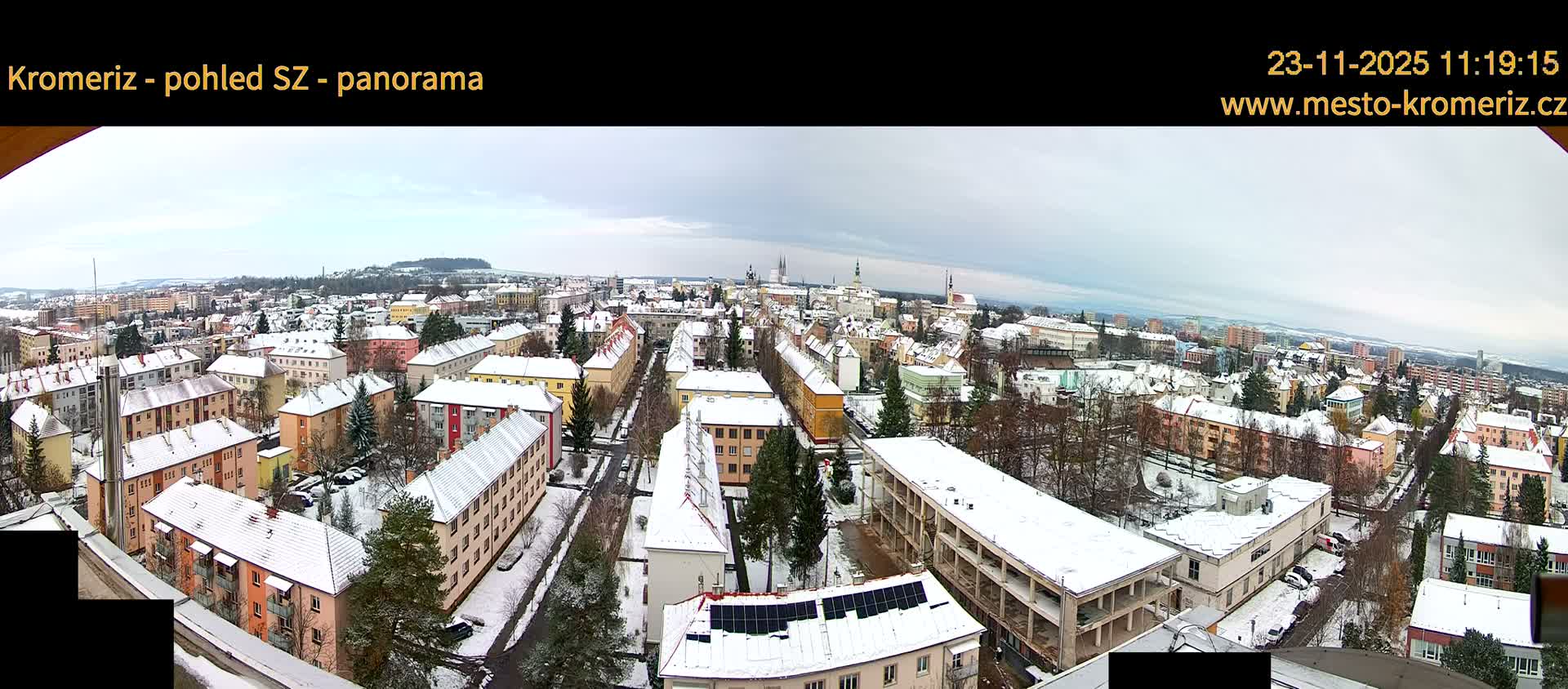 A panoramic view shows a snow-covered townscape with numerous buildings, sparse trees, and streets, all set against distant hills under a cloudy, overcast winter sky.