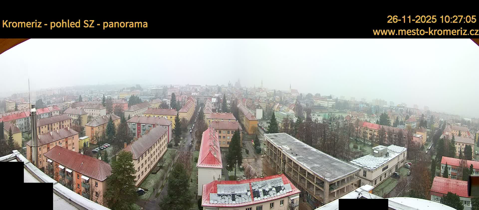 A panoramic view of a European town unfolds on a cold, overcast, and hazy day, with rooftops and sparse winter trees lightly dusted with snow, and distant buildings barely visible through the atmospheric conditions.