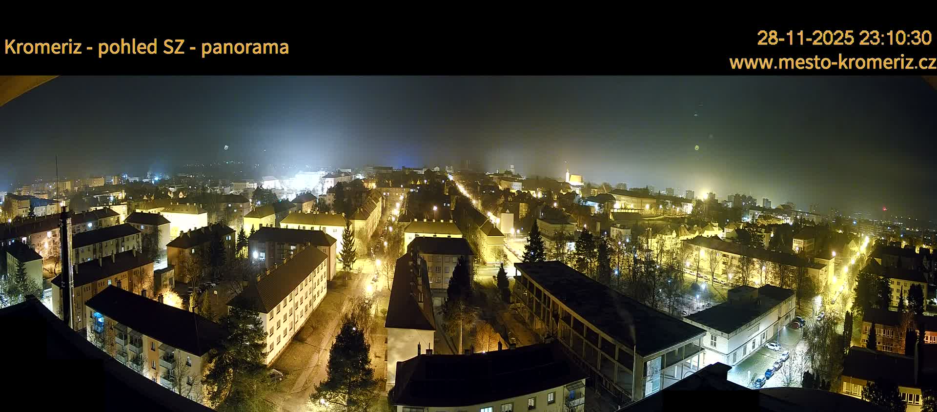 A broad night view reveals a city illuminated by streetlights and building lights under a dark, hazy sky, highlighting residential districts and numerous trees.