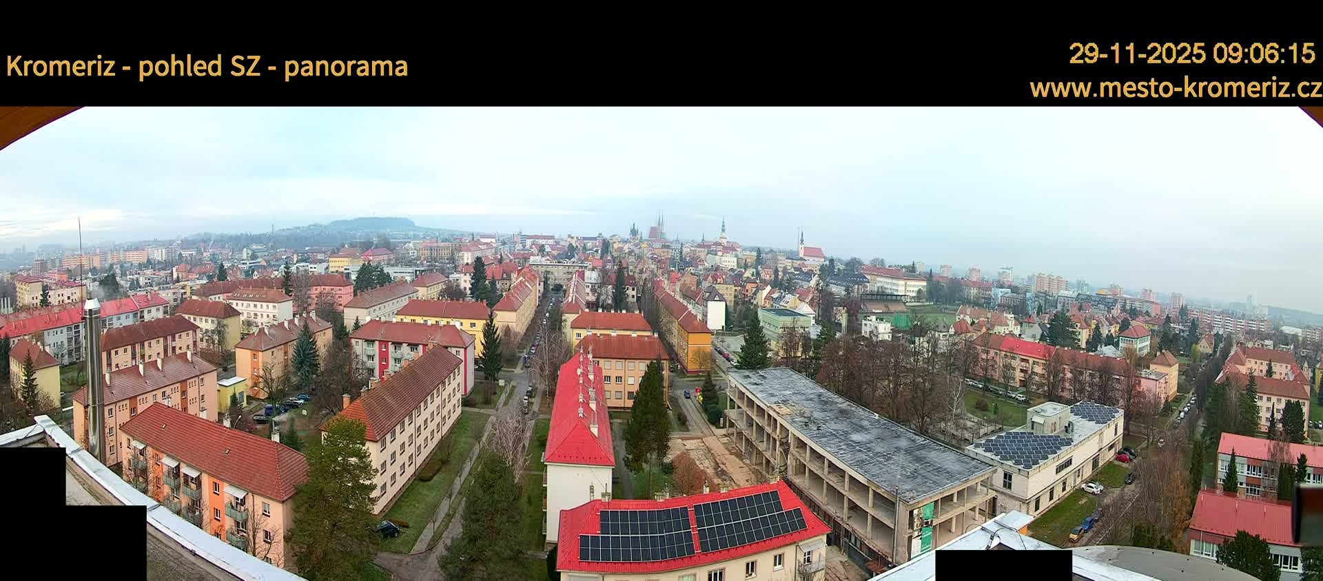A wide panoramic view under an overcast sky depicts a sprawling European town with numerous residential buildings, some featuring red roofs and solar panels, alongside bare trees, an elongated concrete structure, and distant hills, with church spires puncturing the horizon.