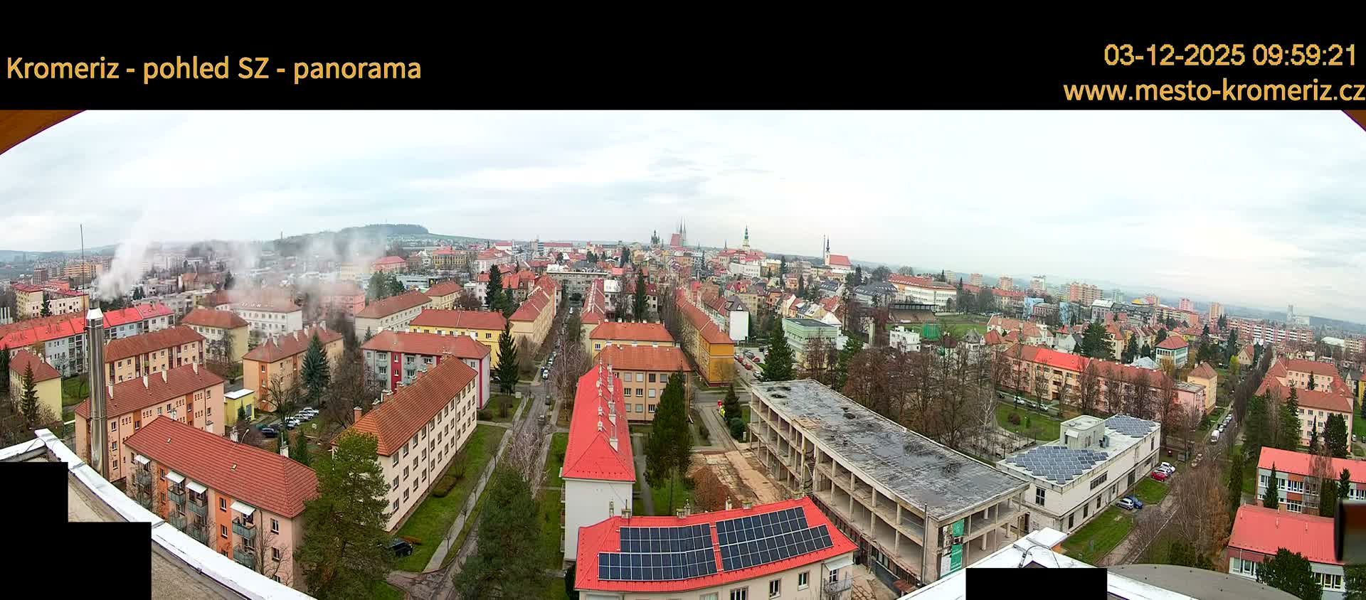 A wide, panoramic view of a sprawling city on an overcast day, featuring a dense mix of residential buildings, prominent church spires in the distance, a large partially constructed building in the foreground, and multiple plumes of smoke rising into the grey sky, with bare trees suggesting a cooler season.